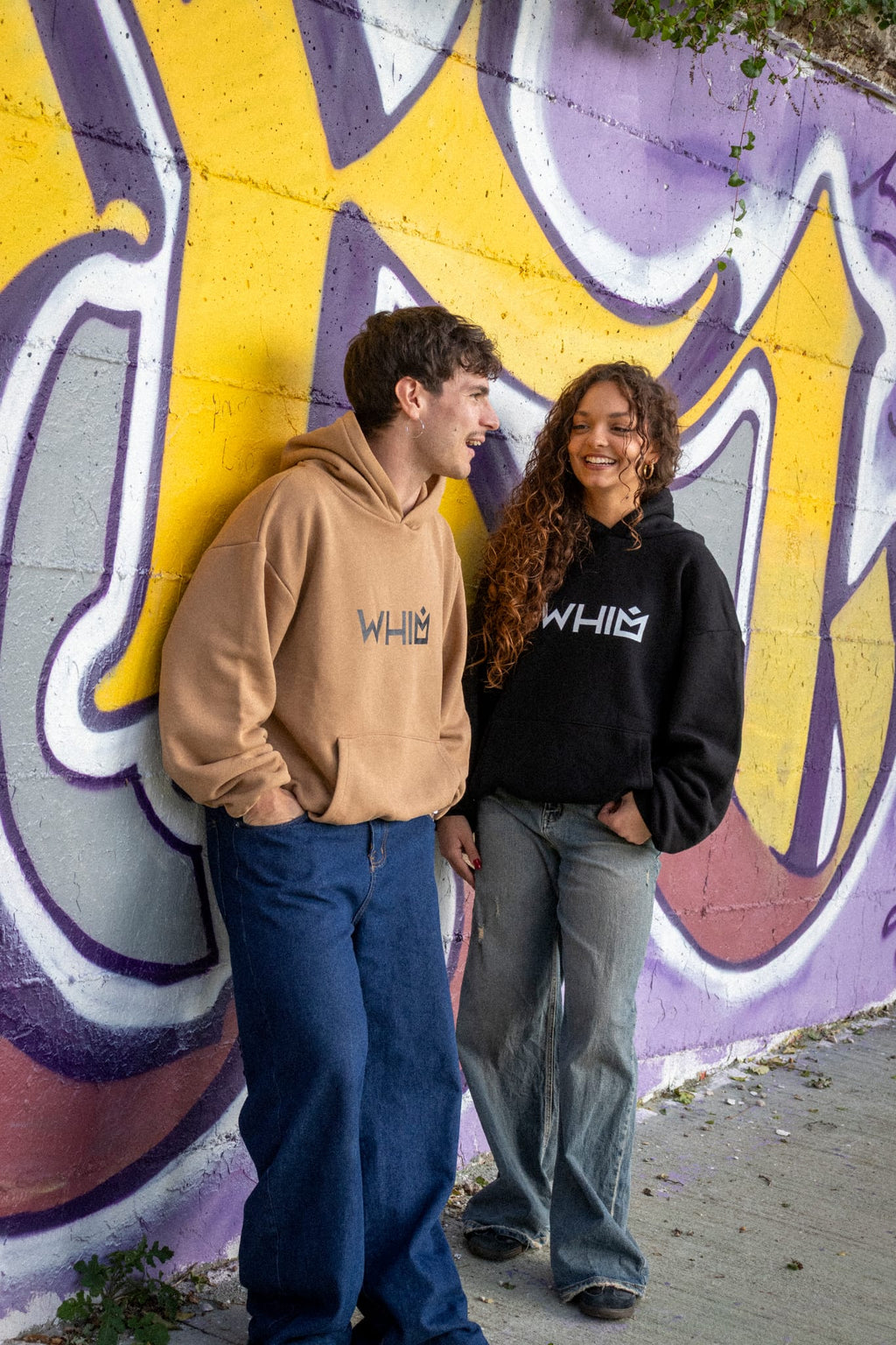 Un modello e una modella indossano felpe unisex WHIM davanti a un muro con murales