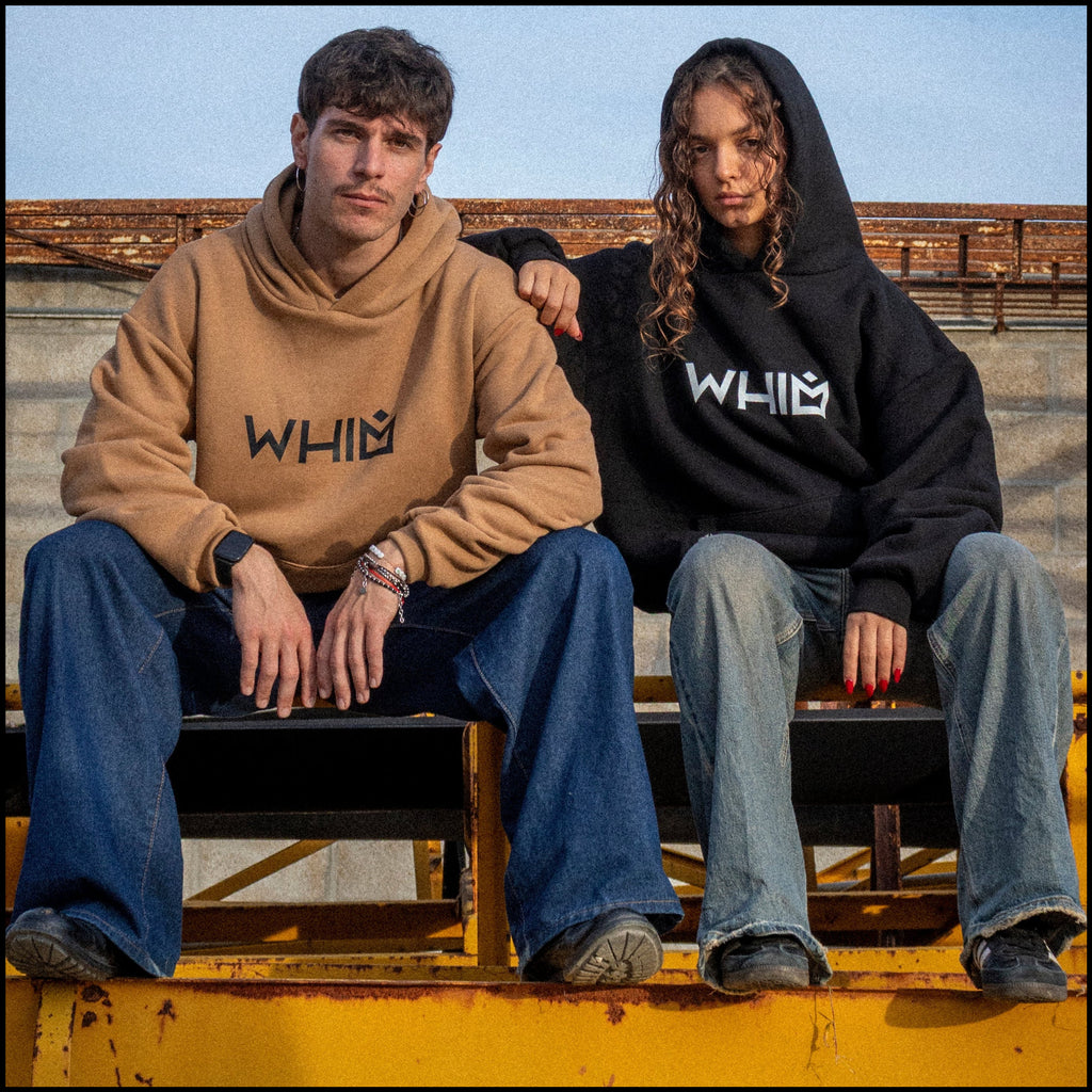 Un modello e una modella seduti su una struttura metallica indossano felpe unisex oversize, una nera e una color cammello.