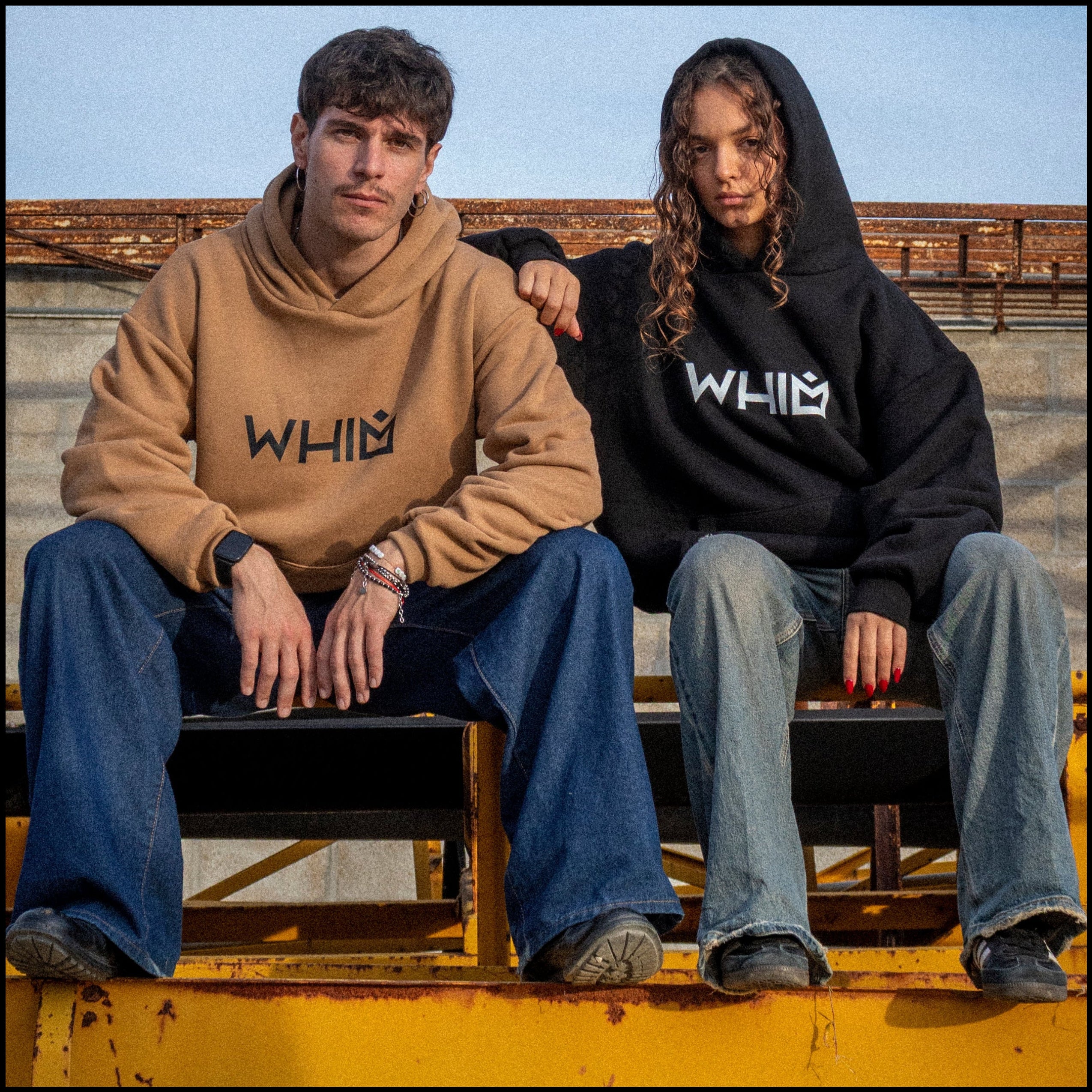 Un modello e una modella seduti su una struttura metallica indossano felpe unisex oversize, una nera e una color cammello.